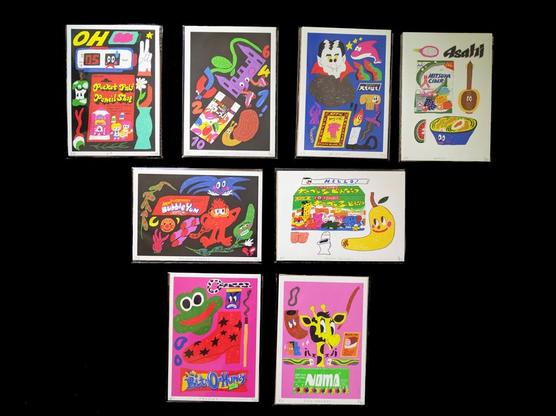 May include: A collection of eight colorful prints featuring various illustrations and text. The prints are framed in black and feature a variety of themes, including food, animals, and abstract designs. The text on the prints includes "Pocket Pal Pencil Sha", "Bubble Yum", "Hillo", "Asahi", "Noma", and "Bite O' Honey".