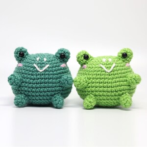 May include: Two crocheted frog plushies, one green and one a darker shade of green, with black eyes and white smiling mouths.