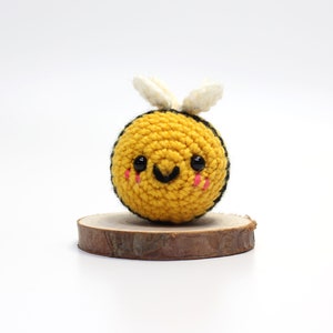 Op de afbeelding: Een gehaakt bij-speelgoed met een geel lichaam, zwarte strepen, witte vleugels en een glimlachend gezicht met zwarte ogen en roze wangen. De bij zit op een houten basis.