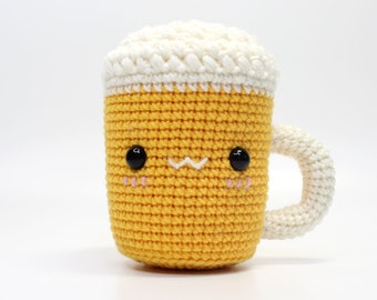 May include: A crocheted beer mug with a white foam top and a cute cartoon face. The mug is yellow and has black eyes and a pink blush.