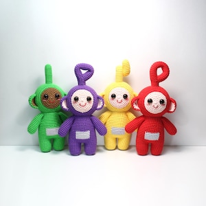 Op de afbeelding: Vier gehaakte poppen in heldere kleuren: groen, paars, geel en rood. Elke pop heeft een lachend gezicht, een gebogen antenne en een witte rechthoek op de torso. De poppen zijn handgemaakt en staan rechtop.