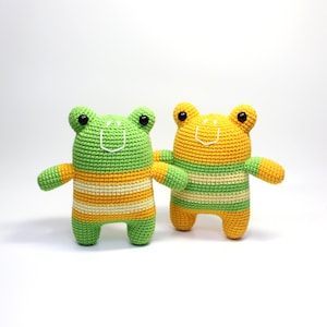 Op de afbeelding: Twee gehaakte knuffels van kikkers, een groene en een gele, met zwarte ogen en een witte glimlachende mond. Beide kikkers hebben een geel-groen gestreept lichaam.