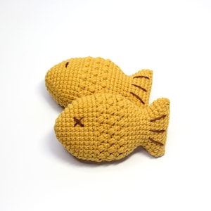 May include: Two crocheted fish-shaped stuffed toys in a yellow color with brown stitching details. The toys are lying on a white surface.