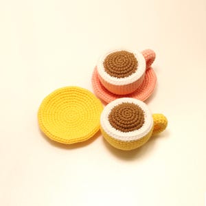 May include: Crocheted coffee cups and saucers in pastel colors. One cup is yellow with a white rim and a brown center, and the other is pink with a white rim and a brown center. A yellow saucer is next to the yellow cup.