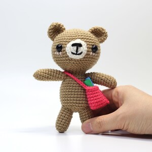 May include: A brown crocheted teddy bear with black eyes and a white nose. The bear is wearing a pink bag with a green and blue strap.