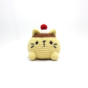 Può includere: Un giocattolo di peluche a forma di gatto all'uncinetto con un corpo giallo, orecchie marroni e un cappello bianco e rosso. Il gatto ha occhi neri e un naso nero.