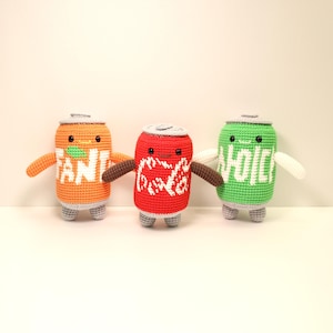 Puede incluir: Tres peluches de ganchillo con forma de lata de refresco. La lata naranja dice "FANTA" en blanco, la lata roja "Coca-Cola" en blanco y la lata verde "VOICE" en blanco. Cada una tiene una tapa plateada, pies grises y brazos.