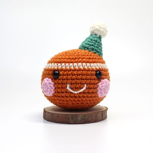 May include: A handmade orange crocheted ball with a smiling face, black eyes, and pink cheeks. It wears a green hat with a white pom-pom. The ball sits on a small wooden slice, creating a whimsical, handcrafted decorative item.