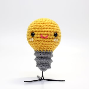 May include: A crocheted lightbulb with a smiling face, yellow body, and gray base. The lightbulb is standing on a black metal stand.