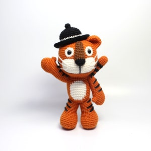 May include: A crocheted tiger in orange and black, wearing a black hat and a white face mask. The tiger has white and brown details, with black stripes on its limbs. It is standing with one arm raised in a waving gesture.
