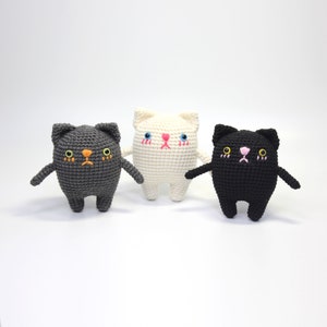 Op de afbeelding: Drie gehaakte katten speelgoedjes, een grijs, een wit en een zwart, met geborduurde gezichtskenmerken. Elke kat heeft een droevige uitdrukking.
