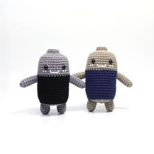 Puede incluir: Dos juguetes de peluche tejidos a crochet con forma de botellas. Uno es gris con la parte superior negra y el otro es marrón con la parte superior azul. Ambos tienen caras sonrientes.