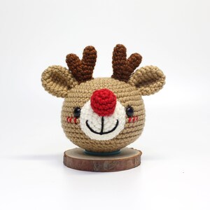 May include: A crocheted reindeer ornament with a brown body, red nose, and black eyes. The reindeer is sitting on a wooden base.