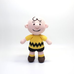 Puede incluir: Un juguete de peluche tejido a crochet de Charlie Brown de la tira cómica Peanuts. El juguete lleva una camisa amarilla con un patrón de zigzag negro y zapatos marrones.