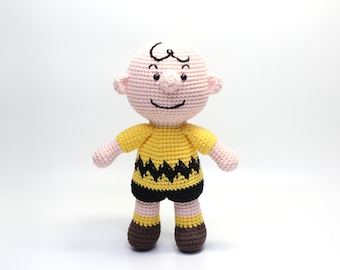 Może przedstawiać: Pluszowa zabawka Charliego Browna z komiksu Peanuts wykonana na szydełku. Zabawka ma na sobie żółtą koszulkę z czarnym wzorem w zygzak i brązowe buty.