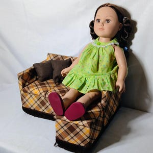 May include: A doll with brown hair and eyes wearing a green dress with a floral pattern and pink shoes, seated on a miniature plaid couch with brown pillows. The couch has a brown, yellow, and white plaid pattern.