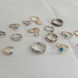 May include: A collection of various rings, including gold, silver, and rose gold tones. Some rings feature gemstones in red and turquoise, while others have unique designs like a heart pendant or a floral motif. The rings are displayed on a white surface.