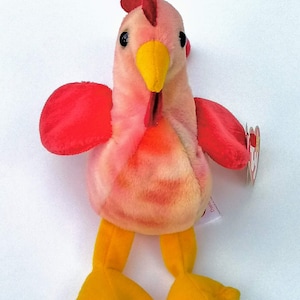 May include: A plush rooster toy in shades of pink, red, and yellow. The rooster has a red comb, wattle, and wings, with a yellow beak and feet. The toy has black eyes and a tag attached to its side.
