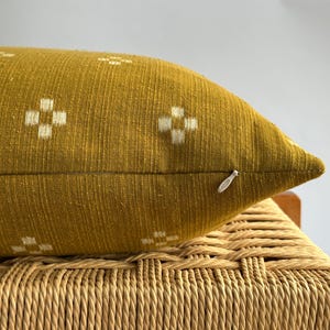 May include: Mustard yellow decorative pillow with a woven texture and white cross-shaped patterns. The pillow is resting on a woven chair. The pillow has a zipper closure.