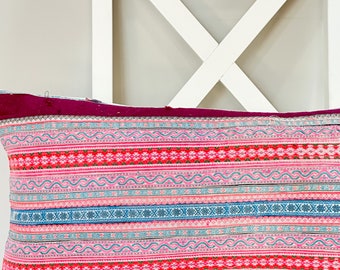 May include: A rectangular decorative pillow with a vibrant, patterned design. The pillow features horizontal stripes in shades of pink, red, and blue, with intricate geometric and floral motifs. The top edge is a deep magenta color.