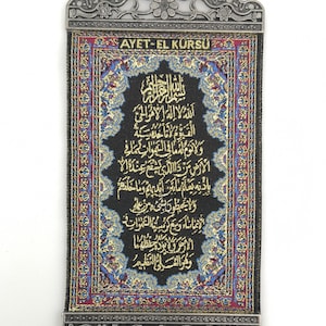 May include: A black fabric wall hanging with gold Arabic calligraphy and a red and blue border. The hanging has a silver metal frame with a loop at the top and silver metal tassels at the bottom. The text on the hanging reads "AYET-EL K&Uuml;RS&Uuml;".