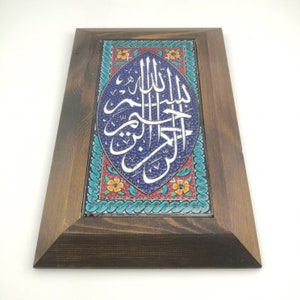 May include: A framed ceramic tile featuring blue background with white Arabic calligraphy. The tile is decorated with a floral border in shades of red, orange, yellow, and green.