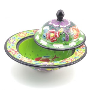 May include: A green ceramic bowl with a lid, both decorated with colourful floral patterns. The bowl has a wide rim and a rounded base. The lid has a knob on top.