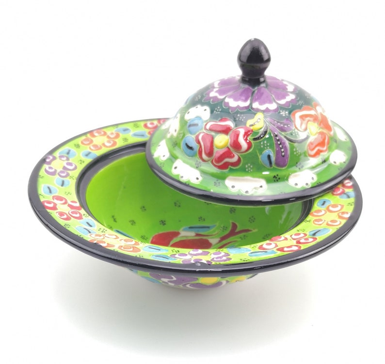 May include: A green ceramic bowl with a lid, both decorated with colourful floral patterns. The bowl has a wide rim and a rounded base. The lid has a knob on top.