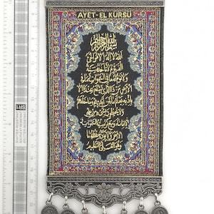 May include: A black fabric wall hanging with gold embroidery featuring Arabic script. The hanging is framed with a silver metal border and has a silver metal loop at the top. The hanging has a silver metal tassel with a silver metal coin at the bottom.