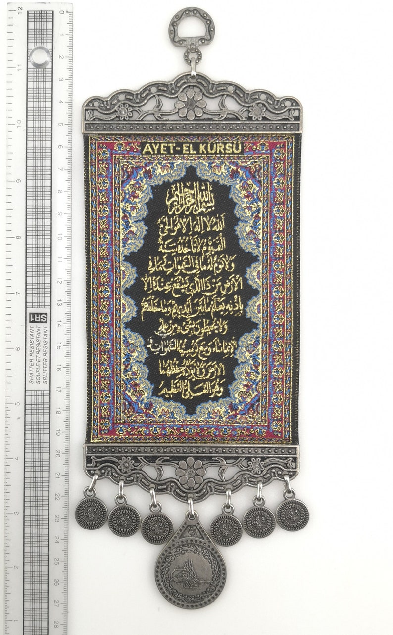 May include: A black fabric wall hanging with gold embroidery featuring Arabic script. The hanging is framed with a silver metal border and has a silver metal loop at the top. The hanging has a silver metal tassel with a silver metal coin at the bottom.