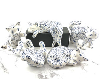 May include: Five ceramic cat figurines with a blue and white floral pattern. The cats are in various poses, including sleeping, sitting, and standing.