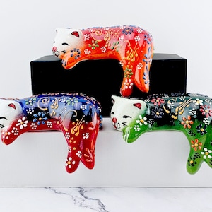 May include: Three ceramic cat figurines in red, blue, and green, each with a white face and intricate floral patterns. The cats are lying down, with their bodies curved. The figurines are decorative and colorful.