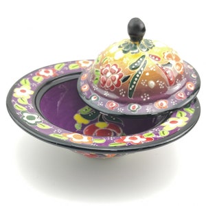 May include: A purple ceramic bowl with a lid, both decorated with colourful floral patterns. The bowl has a wide rim and a rounded base. The lid has a small knob on top.