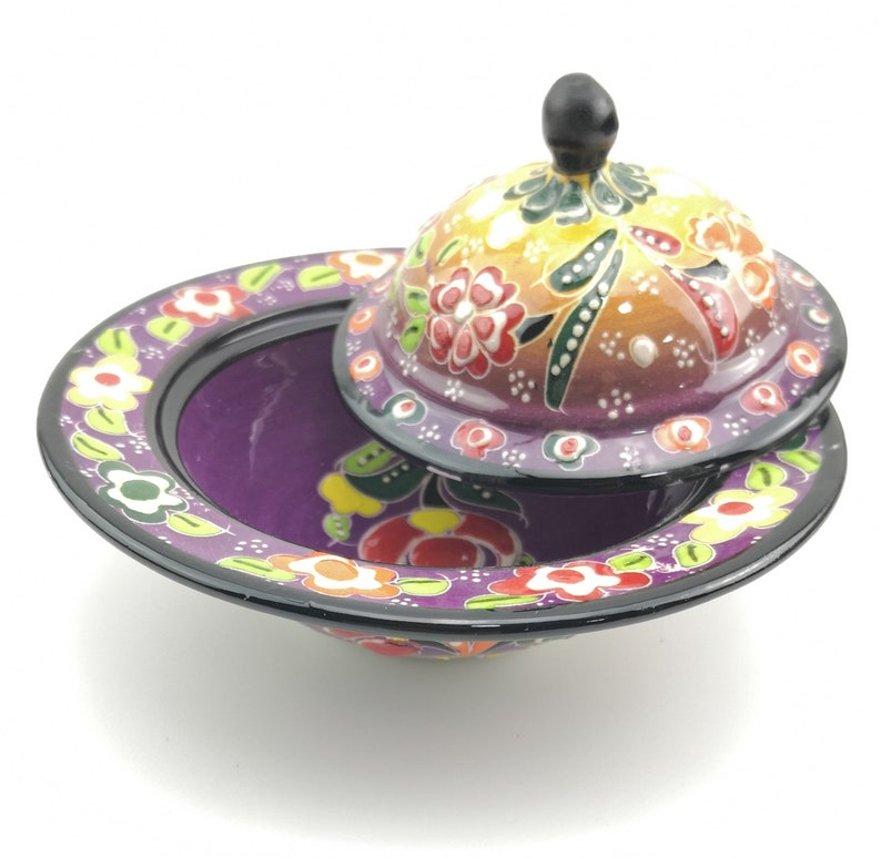 May include: A purple ceramic bowl with a lid, both decorated with colourful floral patterns. The bowl has a wide rim and a rounded base. The lid has a small knob on top.