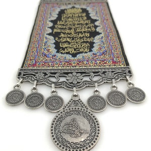May include: A decorative wall hanging with a black background and gold Arabic calligraphy. The hanging is framed with a silver metal frame and has a silver metal tassel with a coin-shaped charm.