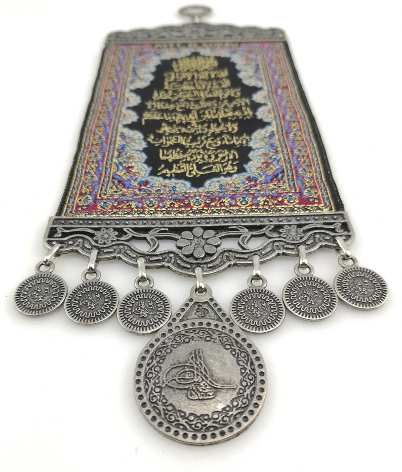 May include: A decorative wall hanging with a black background and gold Arabic calligraphy. The hanging is framed with a silver metal frame and has a silver metal tassel with a coin-shaped charm.