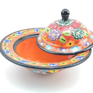 May include: A ceramic bowl with a lid, both decorated with colourful floral patterns. The bowl is orange with black trim, and the lid is red with green, blue, and purple flowers.