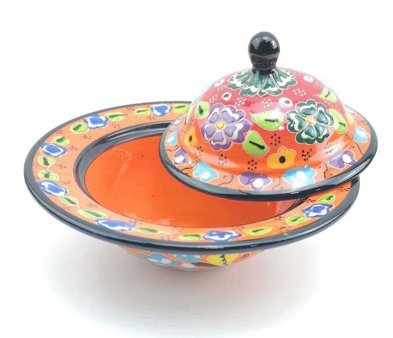May include: A ceramic bowl with a lid, both decorated with colourful floral patterns. The bowl is orange with black trim, and the lid is red with green, blue, and purple flowers.