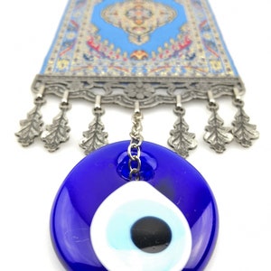 Puede incluir: Un amuleto de ojo turco azul y blanco que cuelga de una cadena plateada. El amuleto est&aacute; unido a una peque&ntilde;a alfombra azul y plateada con flecos.