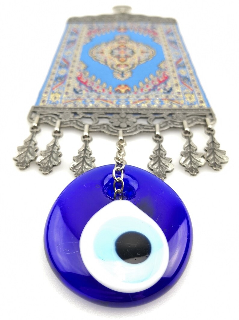 Puede incluir: Un amuleto de ojo turco azul y blanco que cuelga de una cadena plateada. El amuleto est&aacute; unido a una peque&ntilde;a alfombra azul y plateada con flecos.