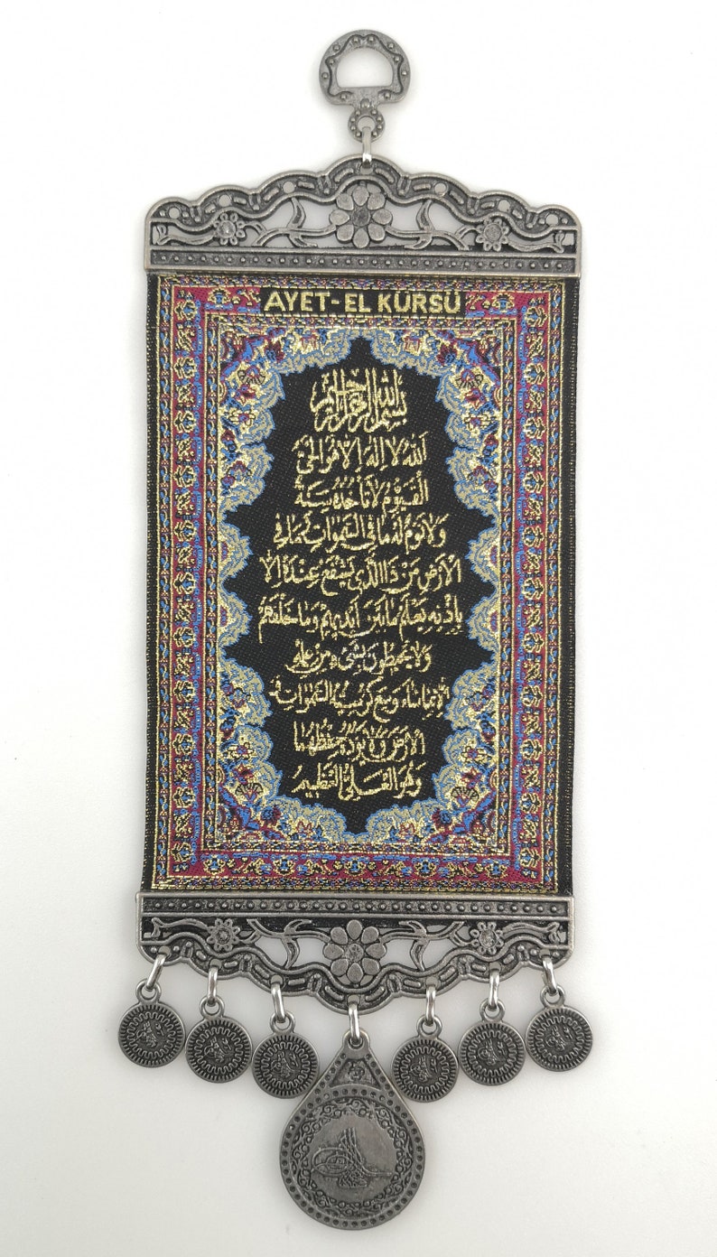 May include: A black fabric wall hanging with gold Arabic calligraphy and a red and blue border. The hanging has a silver metal frame with a loop at the top and silver metal tassels at the bottom. The text on the hanging reads "AYET-EL K&Uuml;RS&Uuml;".