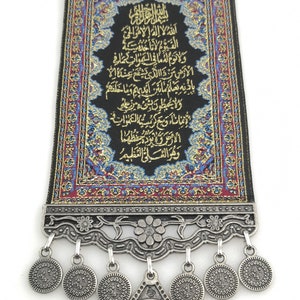 May include: A black fabric wall hanging with gold embroidery featuring the Ayet-el Kursi, a verse from the Quran. The hanging has a silver metal frame with a decorative design and a large silver coin with Arabic calligraphy hanging from the bottom.