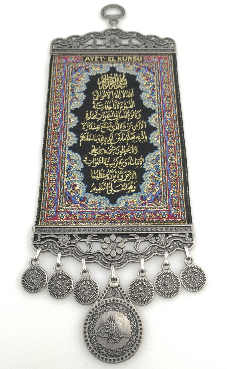 May include: A black fabric wall hanging with gold embroidery featuring the Ayet-el Kursi, a verse from the Quran. The hanging has a silver metal frame with a decorative design and a large silver coin with Arabic calligraphy hanging from the bottom.