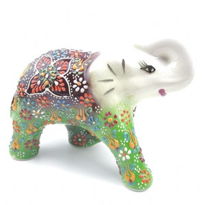 May include: A small, white ceramic elephant figurine with green, brown, and multi-colored floral patterns painted on its body. The elephant has its trunk raised and is standing on all four legs.
