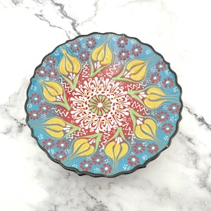 Op de afbeelding: Een blauw en rood keramiek bord met een decoratief ontwerp van gele tulpen en witte bloemen. Het bord heeft een gegolfde rand en een zwarte rand.