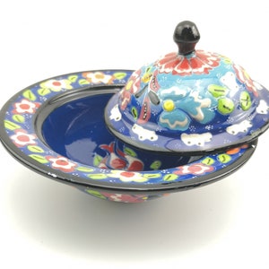May include: A blue and white ceramic bowl with a lid, decorated with colourful floral patterns. The bowl has a wide rim and a rounded base. The lid has a knob on top.