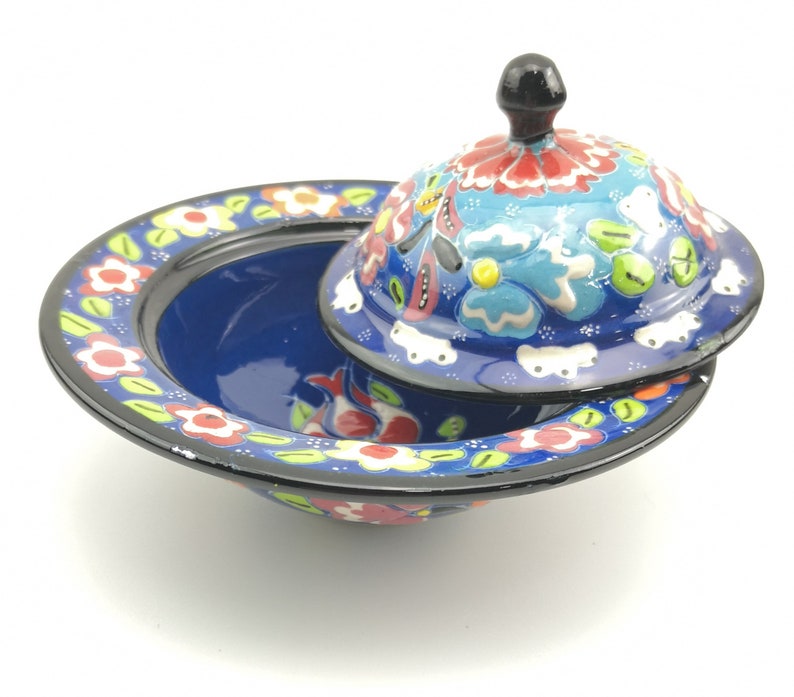 May include: A blue and white ceramic bowl with a lid, decorated with colourful floral patterns. The bowl has a wide rim and a rounded base. The lid has a knob on top.