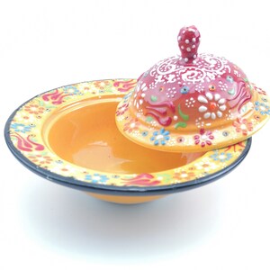 May include: A yellow ceramic bowl with a red and white floral design and a matching lid. The bowl is decorated with colourful flowers and swirls.