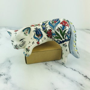 Può includere: Statuetta di gatto in ceramica in posizione di riposo, appoggiata su una scatola di cartone. Il gatto è bianco con intricati motivi floreali in rosso, blu e verde. La coda è azzurra con dettagli gialli. Gli occhi del gatto sono chiusi.