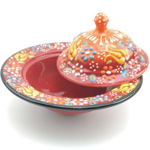 May include: A red ceramic dish with a lid, both decorated with colourful floral patterns and white dots. The lid has a small knob on top.